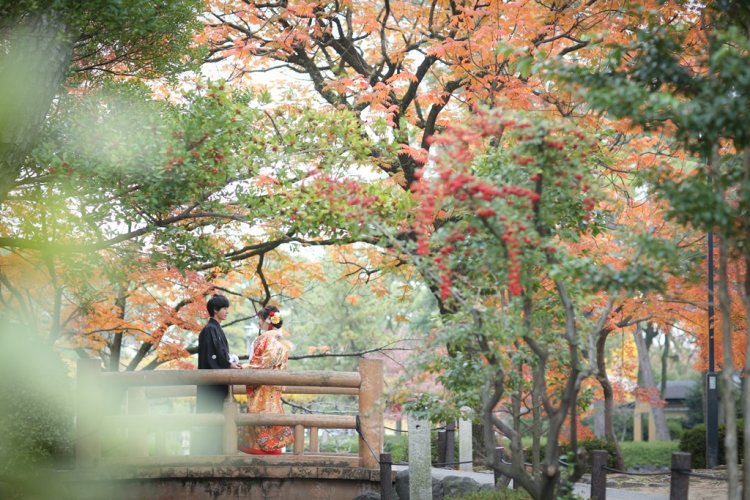 緑の中に色付く紅葉が見られる公園の橋で手を取り合い向かい合う新郎新婦