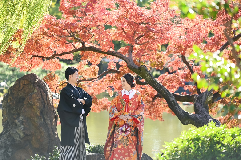 池の前の紅葉の下で見つめ合う笑顔の新郎新婦