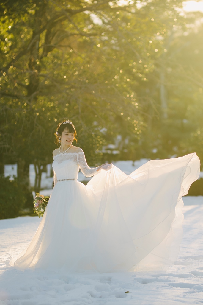 光の射す雪の積もった庭園でドレスをなびかせる新婦